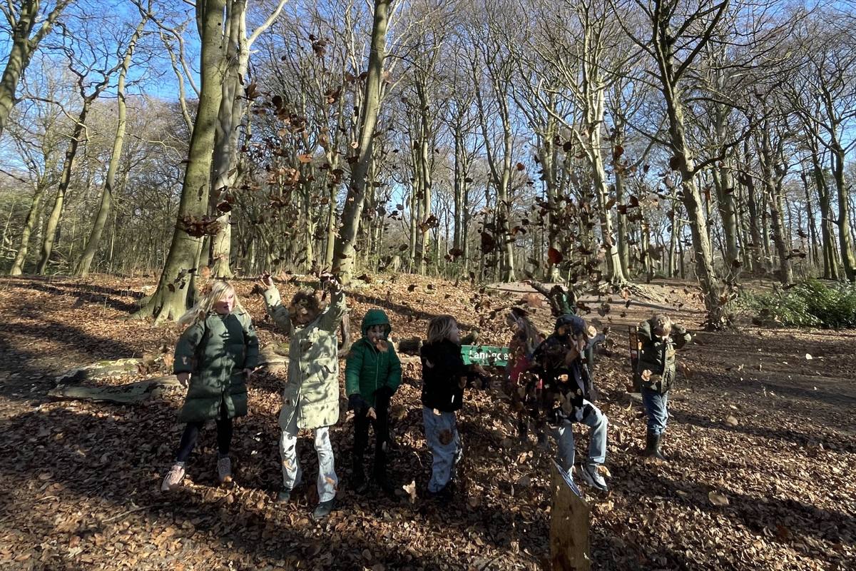 Een groep van zeven kinderen gooit samen een handvol herfstbladeren de lucht in tussen kale bomen