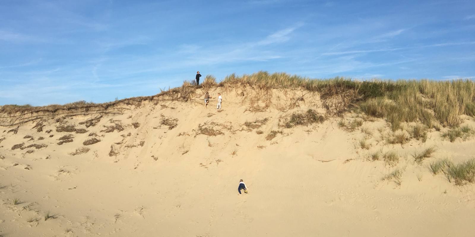 Kleine kinderfiguren tegen een hoge duintop en een wijde lucht, aan de rand van het strand bij Het Schuitengat